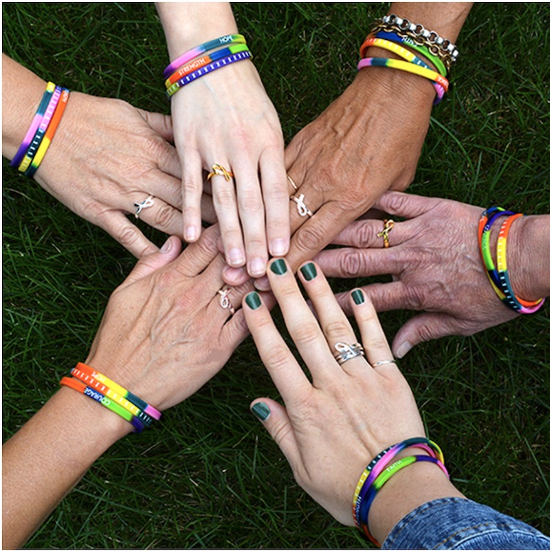 6 hands stacked on top of each other, all wearing rainbow silicone bracelets