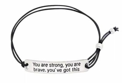 black bracelet with a silver charm reading "You are strong, you are brave, you've got this"