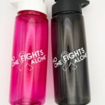 Image showing our No One Fights Alone® Plastic Water Bottle in smoke and in hot pink colors, both with 'No One Fights Alone' and a cancer awareness ribbon printed on the water bottle in white ink.
